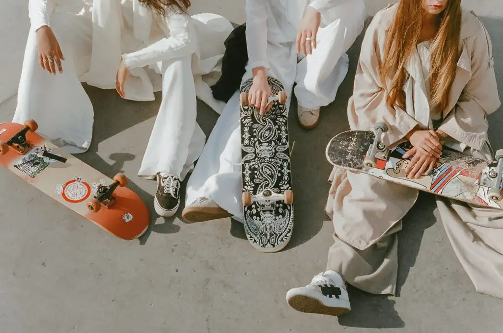 Image of female skateboarders wearing white clothes sitting on the floor while holding skateboards. Source: pexels